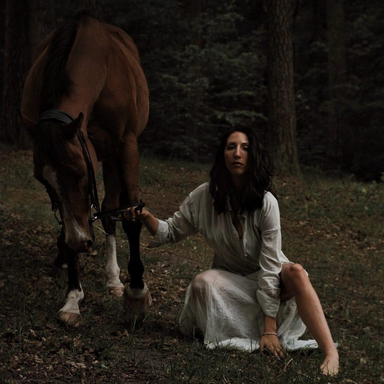 Frau in langem Kleid hockt neben einem Pferd im Wald, Raffaela Richter, Untamed, Ungezähmt, Wildheit, Rückverbindung, Wurzeln, Spüren, Emotionen, Persönlichkeitsentwicklung Frau in weißem Kleid kniet neben einem braunen Pferd im Wald.
