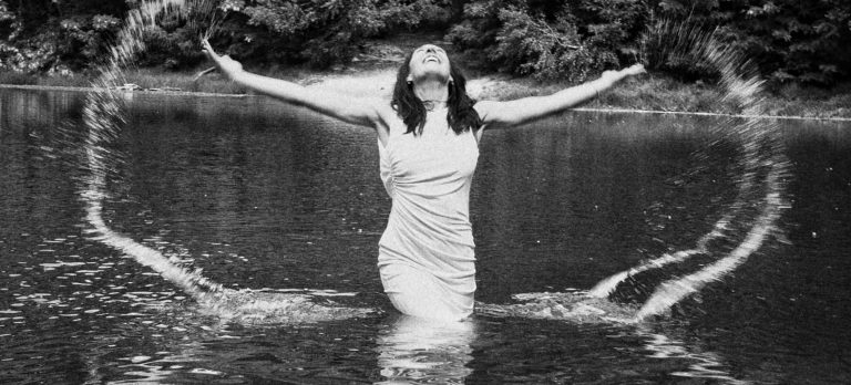 Freie und Fröhliche Frau im Wasser mit ausgebreiteten Armen, Natürlichkeit, Authentizität, Liebe, Freiheit, Gesundheit, ganzheitlich, Breathwork, Coaching, Therapie Eine Frau in einem weißen Kleid steht im Wasser und breitet die Arme aus.